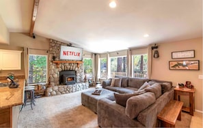 Ein Wohnzimmer mit Kamin aus Stein, einem Netflix-Schild, einer großen Couch und Blick auf Bäume durch die Fenster.