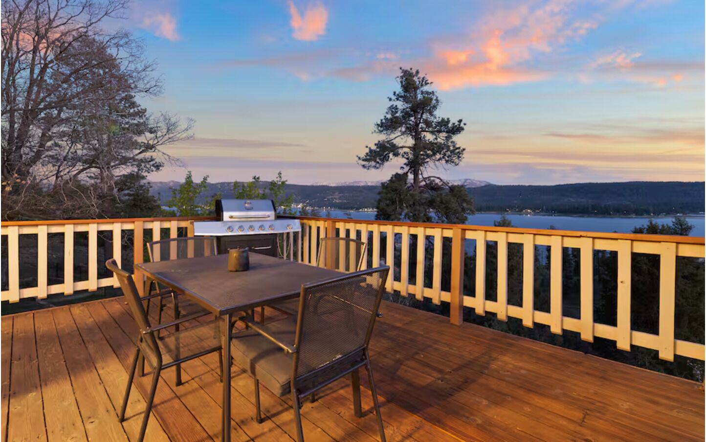 A wooden deck with a grill, outdoor table, and chairs, overlooking a lake and mountains.