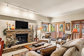 A living room with a stone fireplace, a flat-screen TV, comfortable seating, and a view through a glass door. - On the River, Downtown Ski In/Out Location (Breckenridge)