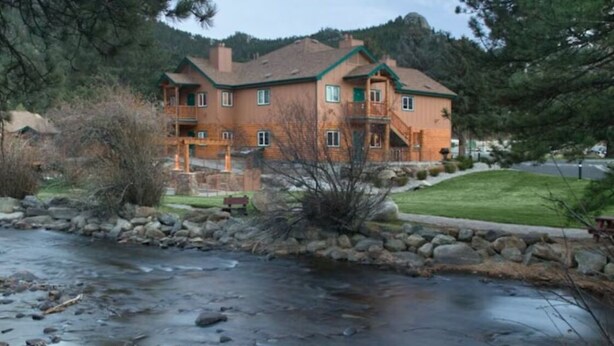 A two-story house with a stone foundation, a wooden deck, and a small stream in front. - Beautiful Riverside 2BR Condo in Estes Park over July 4th, 2026! (Estes Park)
