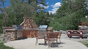 An outdoor patio with stone and metal barbecue grills, a table with chairs, and a picnic table. - Beautiful Riverside 2BR Condo in Estes Park over July 4th, 2026! (Estes Park)