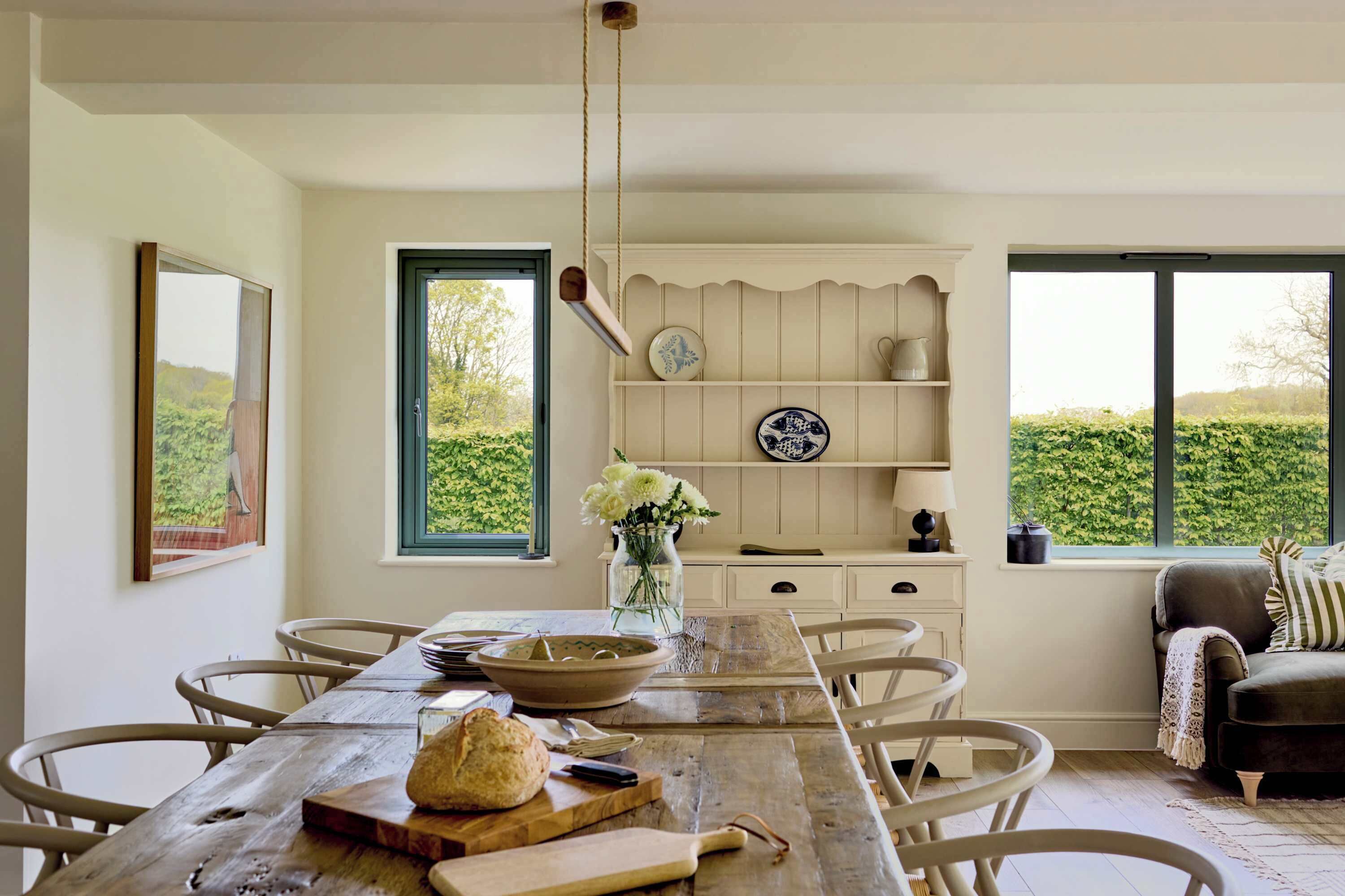 A dining area with a wooden table, chairs, a vase with flowers, and a window with a view of greenery.