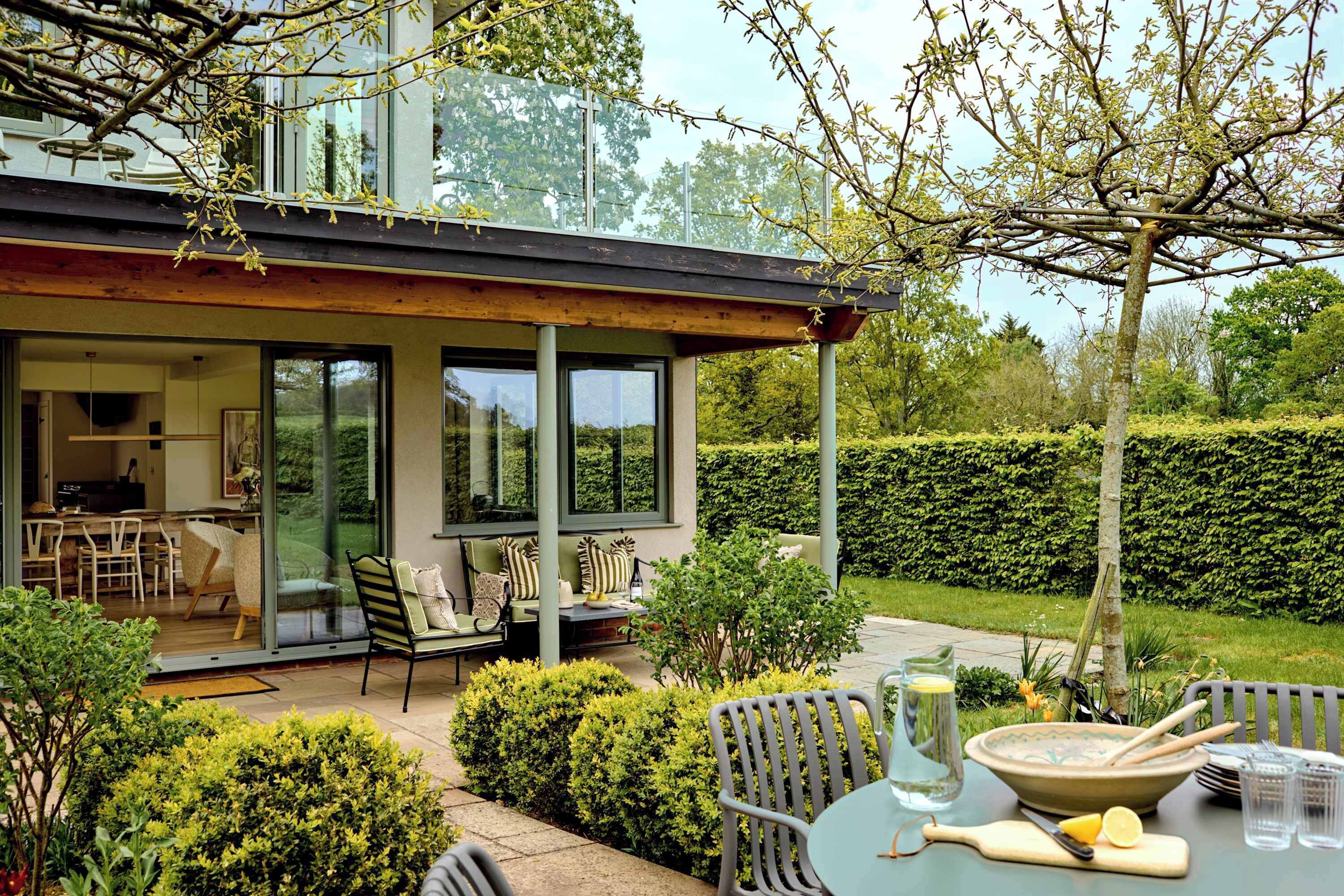 A modern house with a patio area featuring outdoor furniture, a dining table, and a kitchen visible through glass doors.