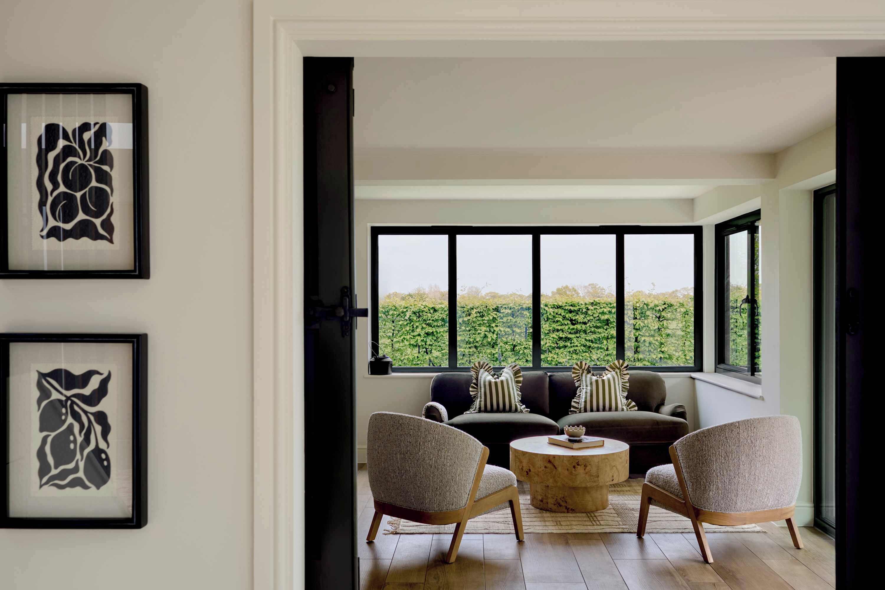 A modern living room with a sofa set, two armchairs, a coffee table, and large windows offering a view of greenery.