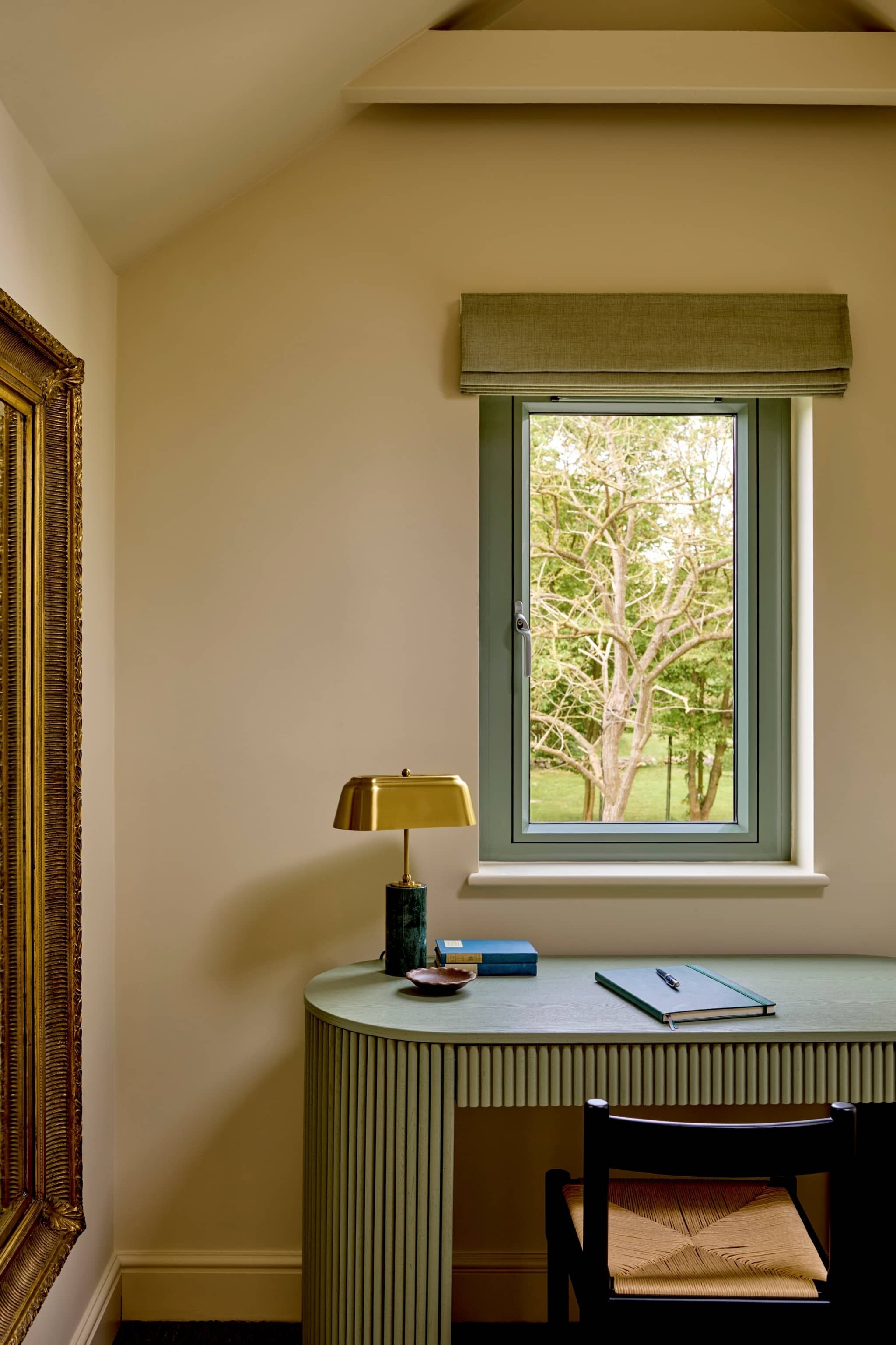 A room with a desk, a chair, a lamp, a window with a view of trees, and a framed picture on the wall.