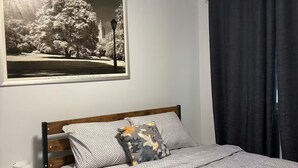Una cama bien hecha con ropa de cama a rayas, un cojín con estampado floral, una lámpara de noche y una fotografía enmarco de un parque.