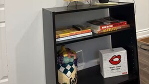 Una habitación con un espejo grande, una televisión, una estantería con libros, una mesita con una planta en maceta y una bolsa de papas.