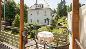Un balcon agrémenté de meubles en osier et d’une table, d’où l’on peut admirer une maison blanche au toit en pente.
