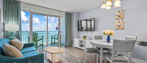 A modern living room with a sofa, dining table, chairs, a TV, artwork, and a balcony with a view.