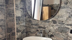 Une salle de bain avec un mur en pierre, un miroir rond, un lavabo blanc, un meuble en bois et un porte-serviettes.