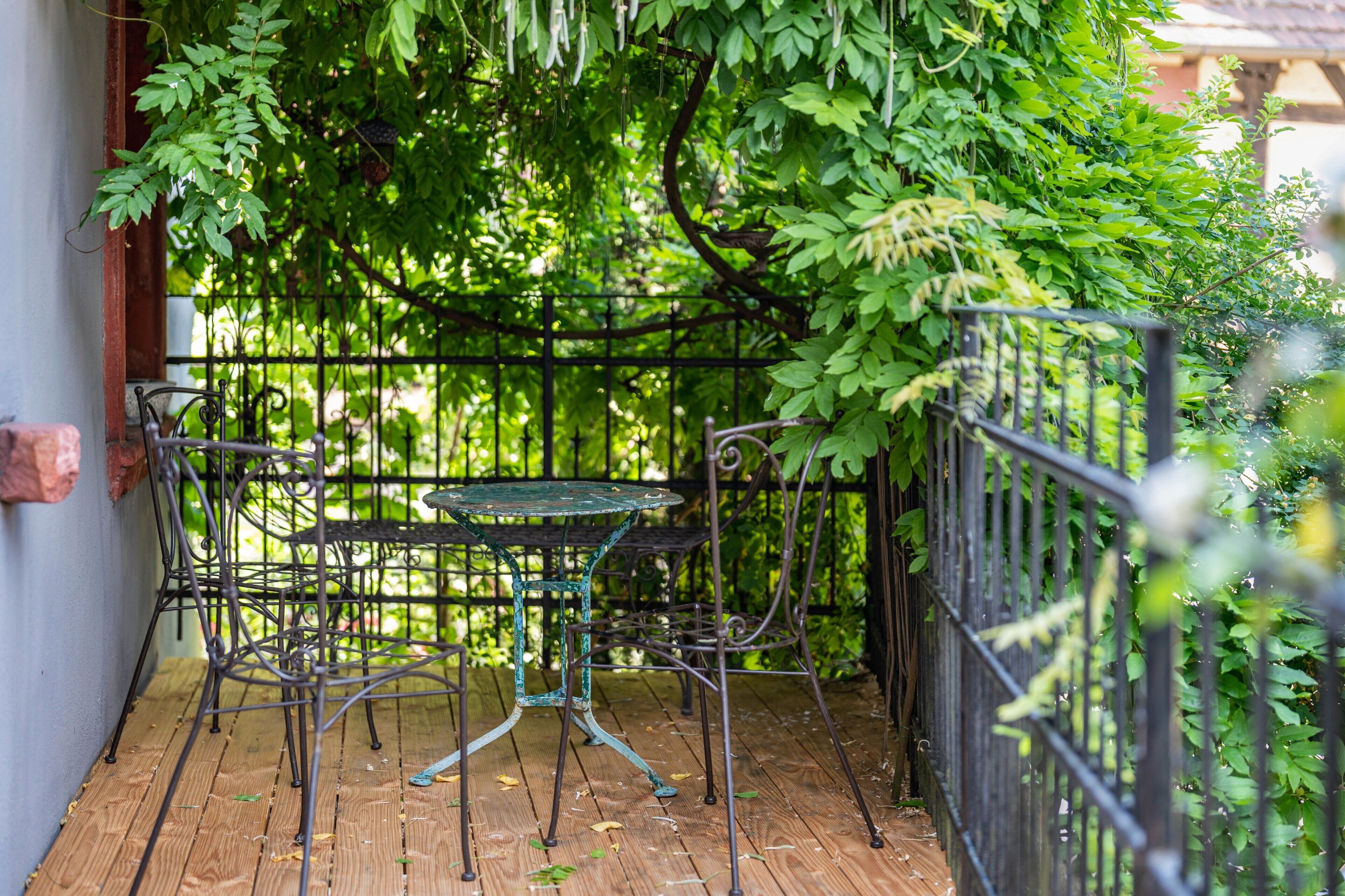 A wooden deck with a metal table and chairs, surrounded by a black metal fence and lush green foliage.