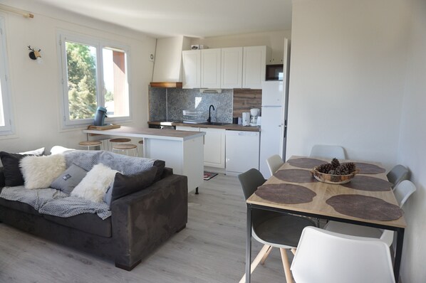Eine moderne Küche mit weißen Schränken, ein Essbereich mit einem Holztisch und Stühlen sowie ein graues Sofa mit Kissen.