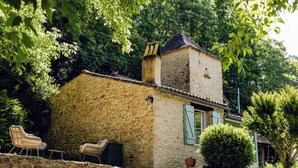 Una casa de piedra con chimenea, una ventana con contraventanas y un pequeño jardín con una planta en maceta y una silla.