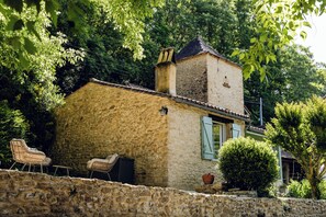 Ein Steinhaus mit Schornstein, einem Fenster mit Fensterläden und einer kleinen Gartenanlage mit Blumentopf und Stuhl.