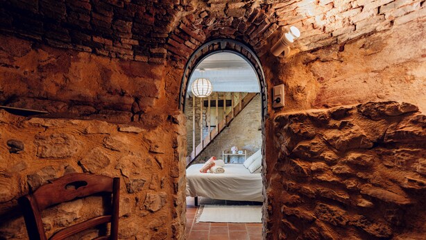 Un estrecho pasillo de piedra que lleva a una cama con colcha blanca, una silla de madera y una escalera al fondo.