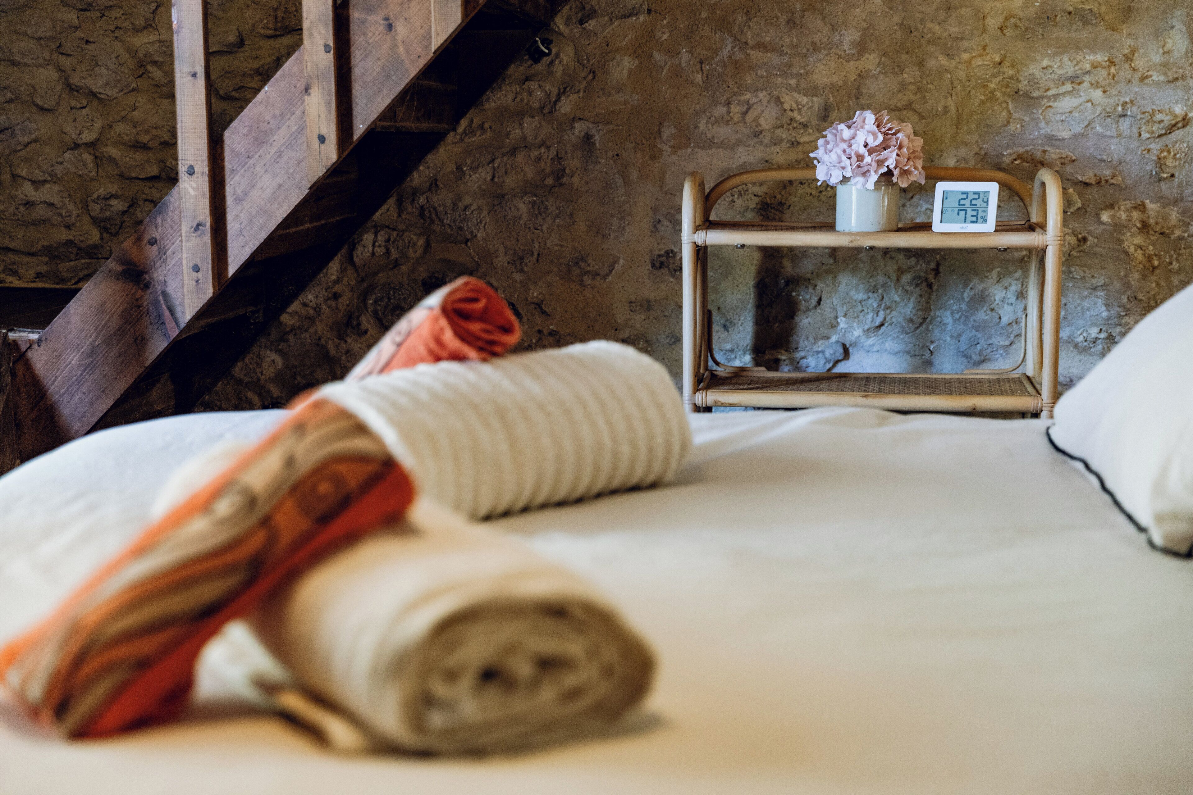 Un lit avec des serviettes enroulées, une table de chevet avec une horloge et des fleurs, et un mur en pierre.