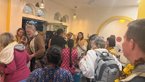 Un espacio interior concurrido, con gente reunida, algunos vistiendo trajes tradicionales y guirnaldas, y un ventilador de techo a la vista.