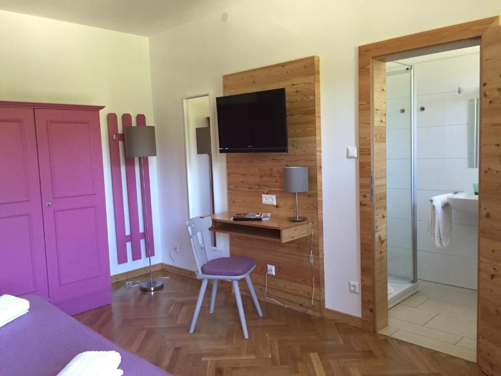 A room with a wooden desk, a chair, a television mounted on the wall, a wardrobe, and a shower area.