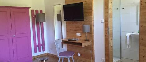 A room with a wooden desk, a chair, a television mounted on the wall, a wardrobe, and a shower area.