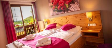 Habitación de hotel con cabecera de madera, cama grande con ropa blanca y detalles rosados, un cuadro de flores en la pared y un balcón con vistas a follaje.