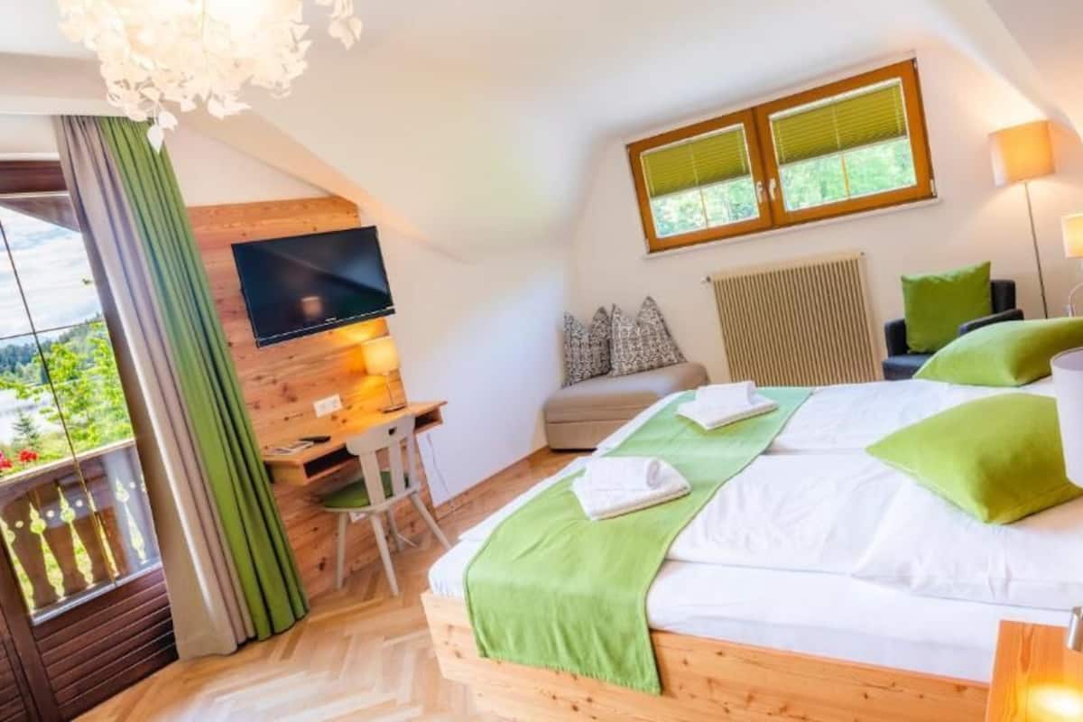 A bedroom with a bed, a TV, a desk, a chair, a window, and a balcony.