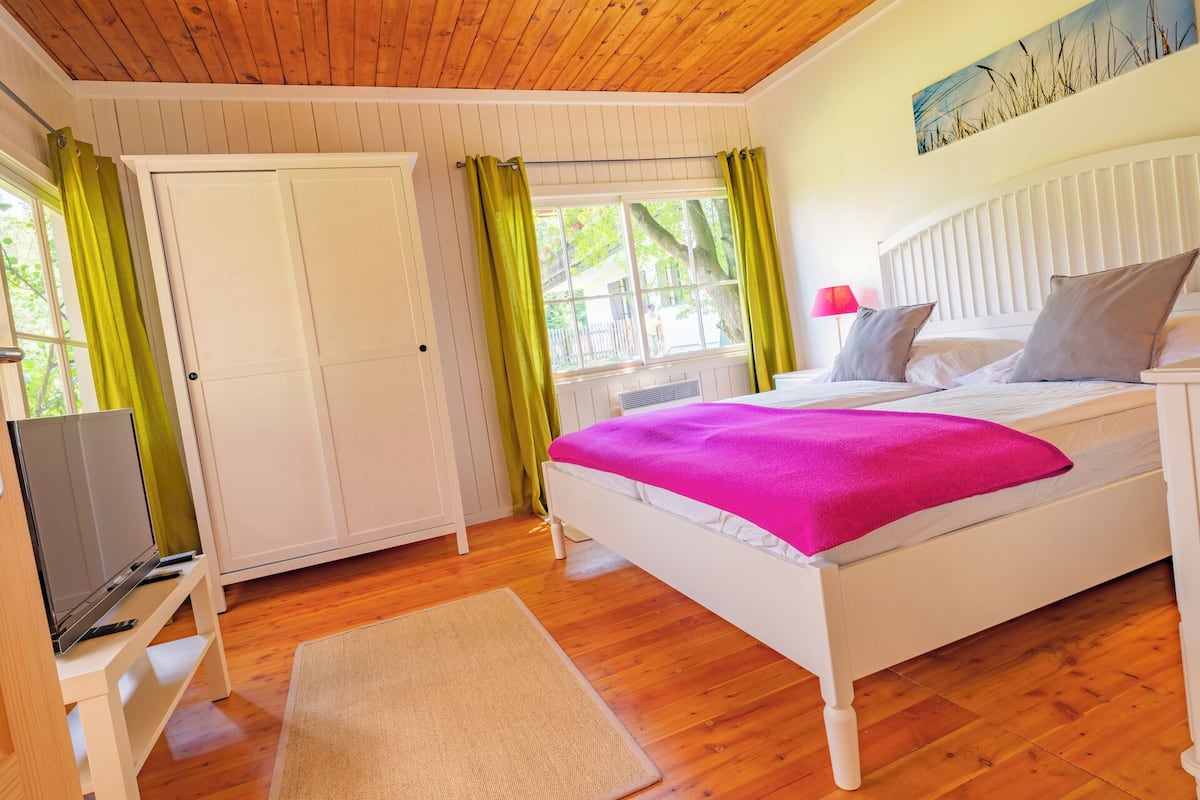 A bedroom with a bed, a television on a stand, a wardrobe, and a window with curtains.