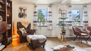 Ein gemütliches Wohnzimmer mit Kamin, Bücherregal, bequemen Sesseln, einem kleinen Tisch mit Pflanze und Blick ins Freie durch große Fenster.