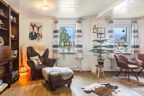 Ein gemütliches Wohnzimmer mit Kamin, Bücherregal, bequemen Sesseln, einem kleinen Tisch mit Pflanze und Blick ins Freie durch große Fenster.