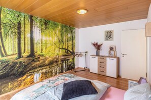 Ein Schlafzimmer mit einem Waldgemälde, Holzboden, einem Bett mit floralem Bettzeug, einem Schminktisch und einer Tür.