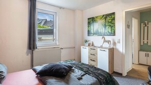 Un dormitori amb un llit, una finestra amb cortines, un radiador, un taulell de nit, un quadre i una porta que condueix a una altra habitació.