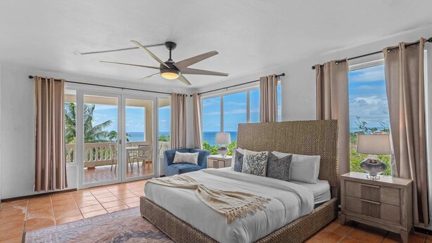 A spacious bedroom with a large bed, a ceiling fan, a nightstand with a lamp, and a view of the ocean through large windows.