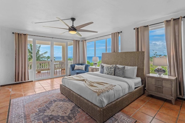 A spacious bedroom with a large bed, a ceiling fan, a nightstand with a lamp, and a view of the ocean through large windows. - Villa Serenidad | Oceanview Pool Retreat (Rincón)