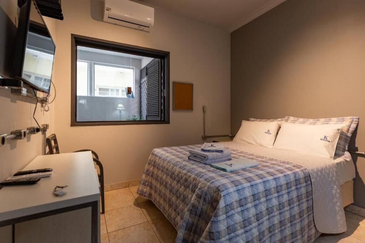 A bedroom with a bed, a desk, a chair, a TV, and a window.