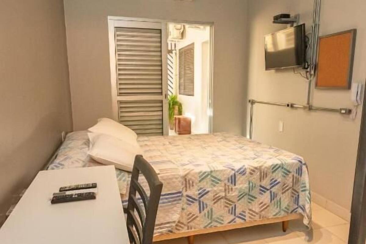 A small, compact room with a bed, a desk, and a chair. There is a television mounted on the wall and an air conditioning unit above the bed.