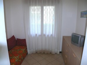 Ein Zimmer mit einem Bett, einem Fernseher auf einer Holzkommode und einem Fenster mit weißen Vorhängen.