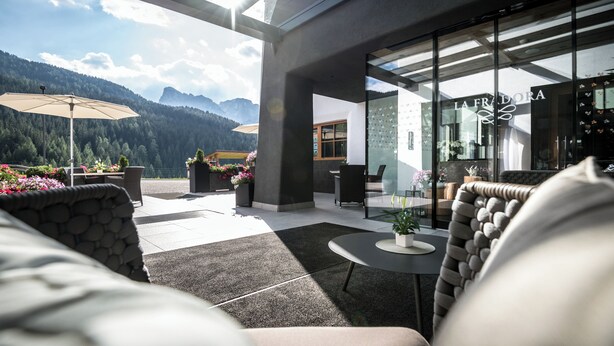 Zona relax esterna moderna con vista sulle montagne, ingresso con porta in vetro e parete in pietra.
