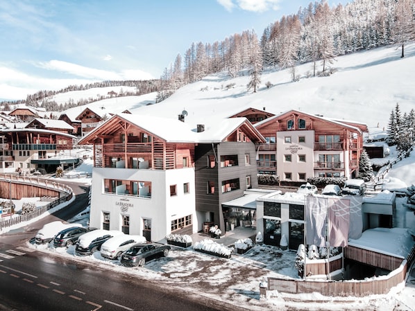 A snow-covered resort with wooden buildings, a parking area, and a ski slope in the background. - Hotel La Fradora - Dolomites Hotel (San Cassiano)