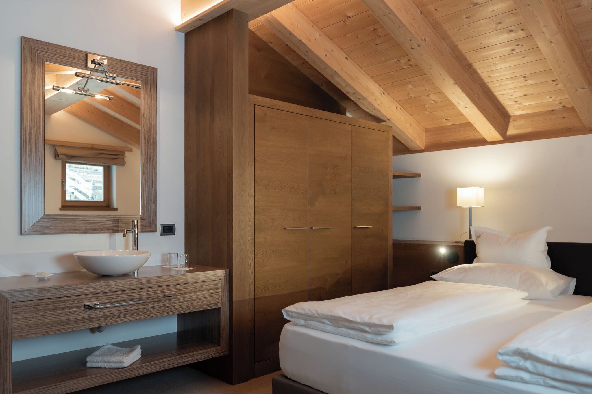 Una camera da letto con soffitto in legno, un armadio grande, un comodino con una lampada e un letto con lenzuola bianche.