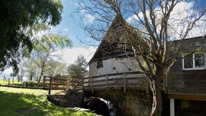 Ein rustikales Steinhaus mit einer Holzbrücke, umgeben von Bäumen und einer Rasenfläche.