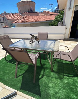 Mesa com tampo de vidro, cadeiras, uma garrafa de vinho e copos em um pátio com grama artificial.