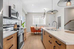A modern kitchen with a central island, a dining area with orange chairs, and a staircase leading to another level. - 2 King Bed | Garage | Min to UofC | NEW (Calgary)