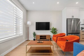 A modern living room with a flat-screen TV, wooden coffee table, orange chairs, and a kitchen area with a stainless steel refrigerator. - 2 King Bed | Garage | Min to UofC | NEW (Calgary)