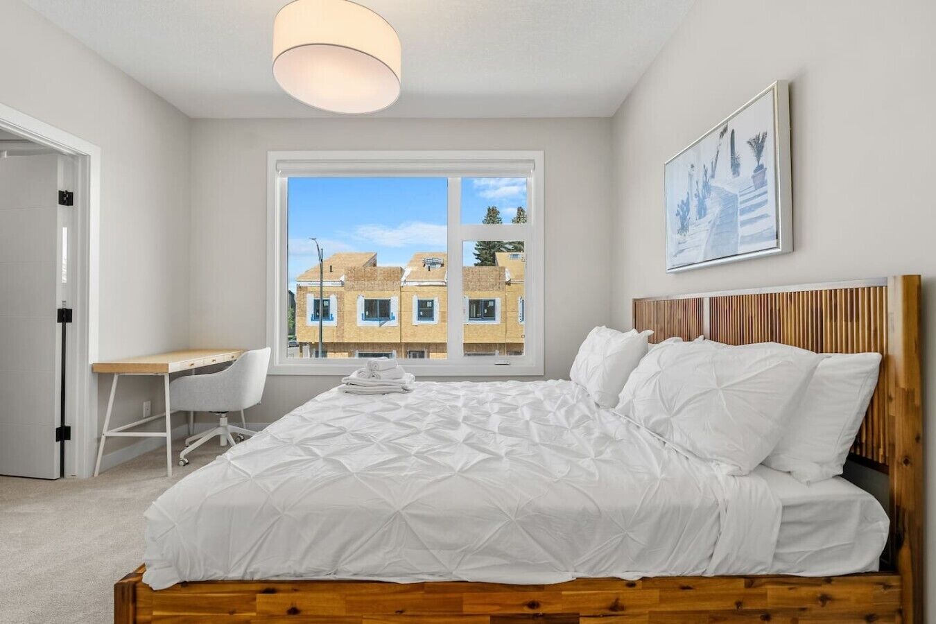 A bedroom with a bed, a desk, and a chair, a window with a view of buildings, and a ceiling light.