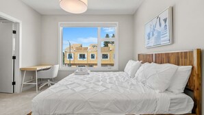 Ein Schlafzimmer mit einem Bett, einem Schreibtisch und einem Sessel, ein Fenster mit Blick auf Gebäude und einer Deckenleuchte.