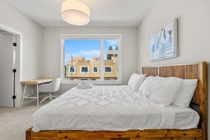Ein Schlafzimmer mit einem Bett, einem Schreibtisch und einem Sessel, ein Fenster mit Blick auf Gebäude und einer Deckenleuchte.