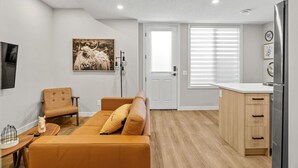 Ein modernes Wohnzimmer mit einer Couch, einem Sessel, einem kleinen Tisch mit einer Dekoration, einem aufgehängten Bild und einer Küchenzeile mit einem Kühlschrank.