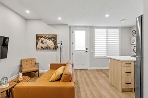 Ein modernes Wohnzimmer mit einer Couch, einem Sessel, einem kleinen Tisch mit einer Dekoration, einem aufgehängten Bild und einer Küchenzeile mit einem Kühlschrank.