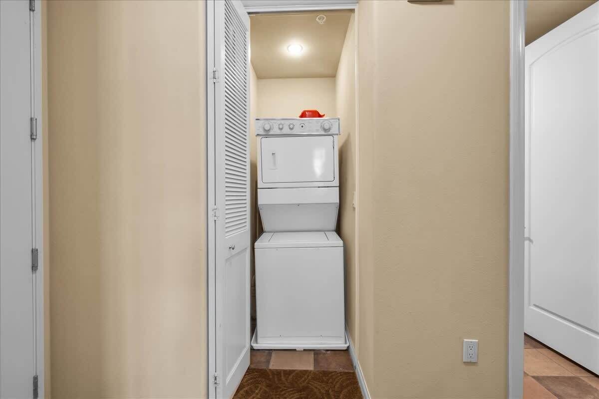 A laundry room with a stacked washer and dryer, a red object on top of the dryer, and a door leading to another room.