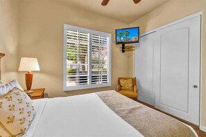 Ein Hotelzimmer mit Bett, Wandfernseher, Fenster mit Jalousien, Lampe und Sessel.