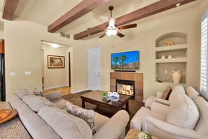A living room with a fireplace, a ceiling fan, and a television. - Desert Serenity #A - 1 BR Legacy Villas (La Quinta)